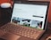 A laptop with a keyboard is open on a desk displaying a webpage about remote work. The screen shows images and text related to working from home, with tabs visible at the top of the browser. There is a smart speaker to the left and part of a chair visible at the edge of the frame.