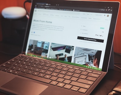 A laptop with a keyboard is open on a desk displaying a webpage about remote work. The screen shows images and text related to working from home, with tabs visible at the top of the browser. There is a smart speaker to the left and part of a chair visible at the edge of the frame.