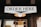 A wooden sign with bold black letters reading 'ORDER HERE' is prominently displayed. Below the sign, a warm glowing lamp adds an inviting ambiance. The setting appears to be part of a rustic exterior, possibly a coffee shop or restaurant.