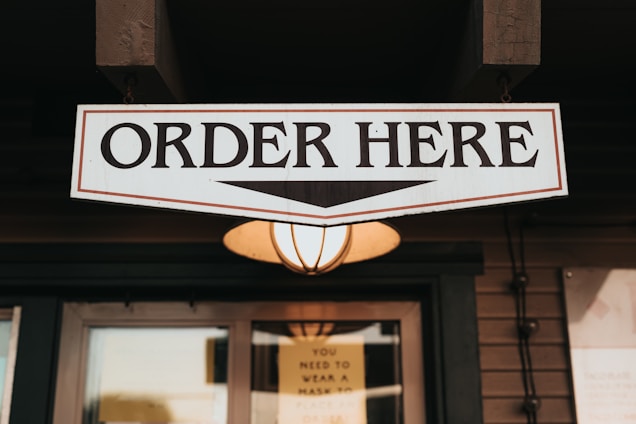 A wooden sign with bold black letters reading 'ORDER HERE' is prominently displayed. Below the sign, a warm glowing lamp adds an inviting ambiance. The setting appears to be part of a rustic exterior, possibly a coffee shop or restaurant.