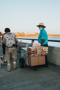 A man wearing a hat and a blue shirt, also wearing a face mask, stands beside a cart filled with various snacks such as chips and candy. The snack cart is positioned on a waterfront walkway with a backdrop of calm water and a distant shore. Another person, partially visible, is holding a small dog on a leash.