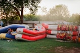 Several inflatable rafts are stacked on a grassy area near a tree. Two children are playing on them, with one child sitting on a blue raft and another climbing on a red one. A rack with numerous life jackets is positioned to the right, adding a sense of readiness for water activities. The setting appears to be outdoors, possibly in a backyard or recreational area, with a fence and trees in the background.