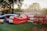 Several inflatable rafts are stacked on a grassy area near a tree. Two children are playing on them, with one child sitting on a blue raft and another climbing on a red one. A rack with numerous life jackets is positioned to the right, adding a sense of readiness for water activities. The setting appears to be outdoors, possibly in a backyard or recreational area, with a fence and trees in the background.
