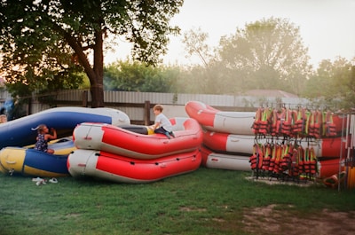 Several inflatable rafts are stacked on a grassy area near a tree. Two children are playing on them, with one child sitting on a blue raft and another climbing on a red one. A rack with numerous life jackets is positioned to the right, adding a sense of readiness for water activities. The setting appears to be outdoors, possibly in a backyard or recreational area, with a fence and trees in the background.