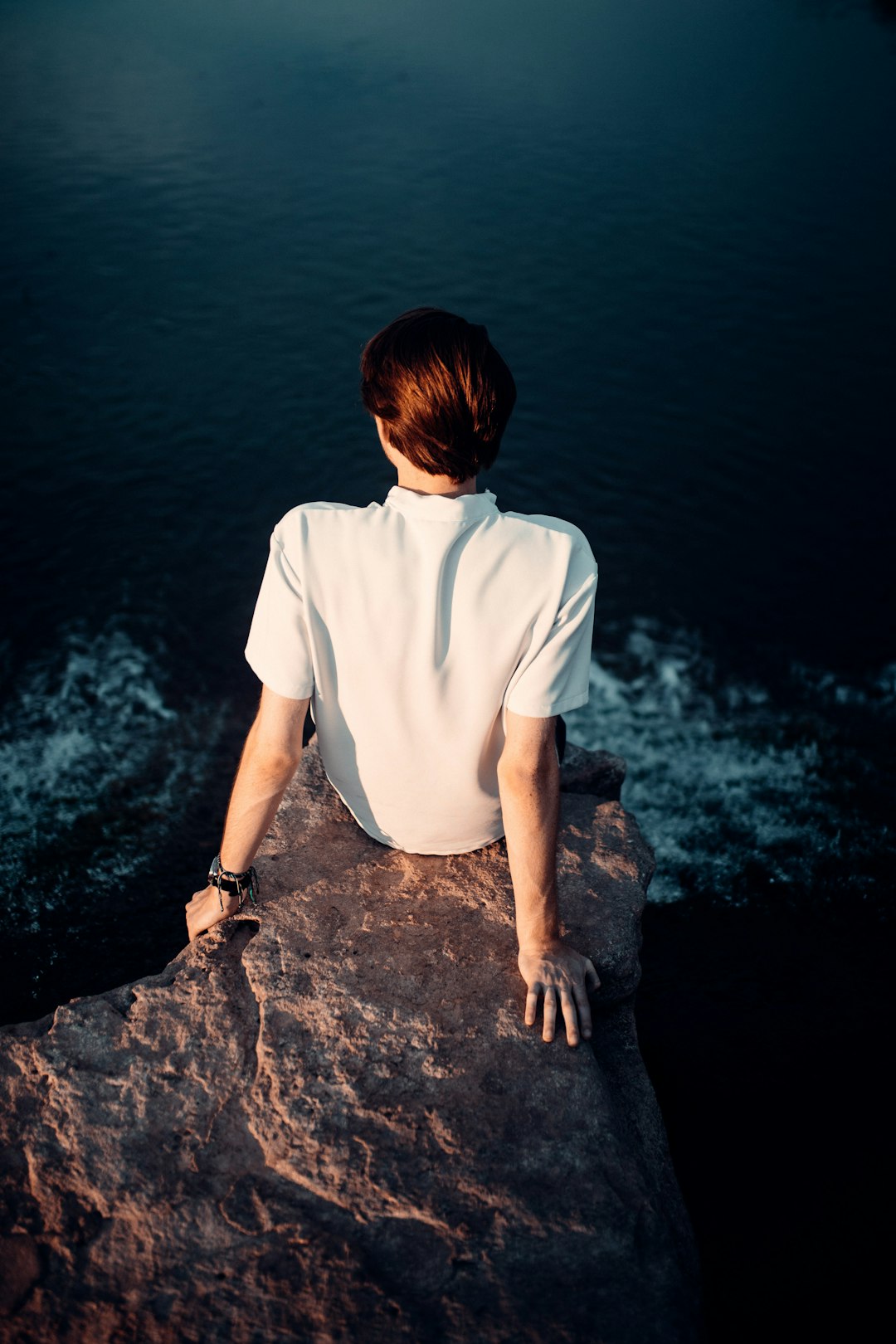 man in white crew neck t-shirt sitting on rock near body of water during daytime