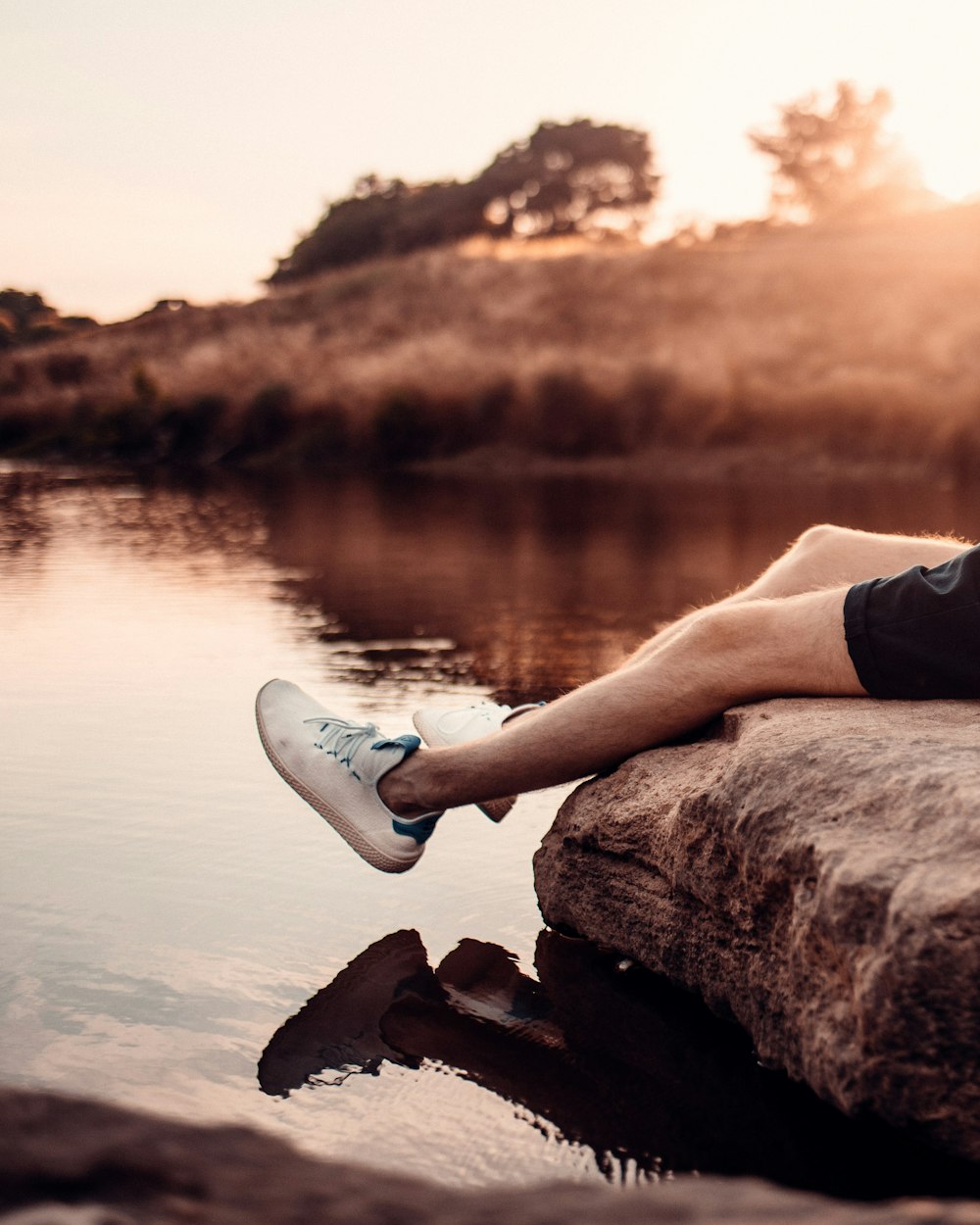 person wearing white sneakers sitting on brown rock near body of water during sunset