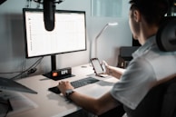 A person is seated at a desk using a computer monitor and holding a smartphone. The monitor displays a spreadsheet or data sheet, and the desk is equipped with a keyboard, a digital clock displaying the time 6:34, and a desk lamp.