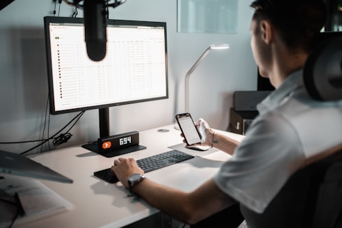 A person is seated at a desk using a computer monitor and holding a smartphone. The monitor displays a spreadsheet or data sheet, and the desk is equipped with a keyboard, a digital clock displaying the time 6:34, and a desk lamp.
