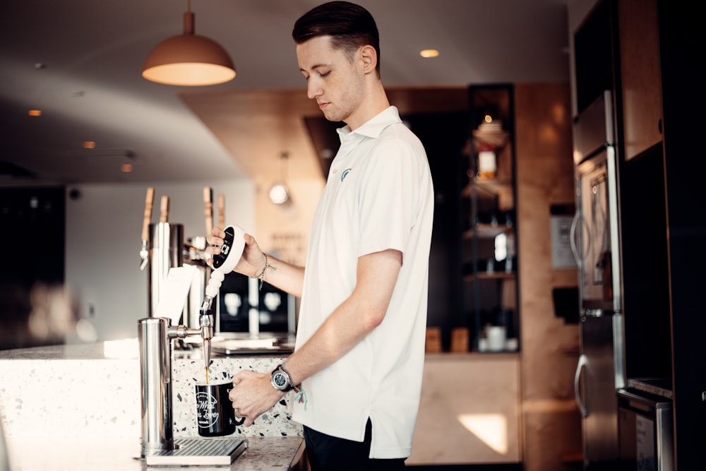 homem de botão branco até camiseta e calças pretas em pé na frente da máquina de café expresso