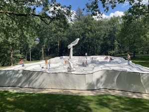 A group of people interacts with a large sculpture resembling a black and white comic book drawing. The artwork features bold outlines and abstract shapes on a raised platform surrounded by a lush green forest.