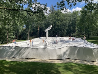 A group of people interacts with a large sculpture resembling a black and white comic book drawing. The artwork features bold outlines and abstract shapes on a raised platform surrounded by a lush green forest.