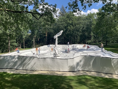A group of people interacts with a large sculpture resembling a black and white comic book drawing. The artwork features bold outlines and abstract shapes on a raised platform surrounded by a lush green forest.