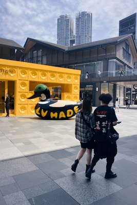 A large black and yellow inflatable duck with 'LV MADE' written on it is displayed outside a building with a speaker-inspired facade. Two people are walking in the foreground, and tall buildings are visible in the background. The setting appears to be a modern urban shopping area with clear skies.