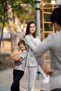 A young woman and a boy stand back-to-back with arms crossed in an outdoor setting. The boy leans against the woman with a playful and confident expression. Trees and playground equipment are visible in the background.