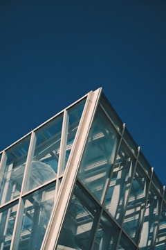 A modern architectural structure made of glass panels with a visible steel frame, set against a clear blue sky. The building's design includes multiple large transparent panels that reflect light and reveal some internal mechanisms.