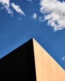 A minimalist perspective of a building corner with a sharp angle, contrasting against a clear blue sky with a few scattered clouds.