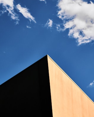 A minimalist perspective of a building corner with a sharp angle, contrasting against a clear blue sky with a few scattered clouds.
