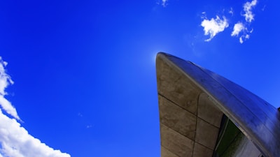A large, angled concrete structure extends upwards into a bright blue sky. Fluffy white clouds are scattered across the sky, adding contrast. The structure appears modern and industrial, with smooth surfaces and sharp lines.