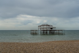 A desolate scene featuring the rusted, skeletal remains of a pier extending into the sea, with a cloudy sky above and a pebbled beach in the foreground. The structure appears abandoned and weathered by time.