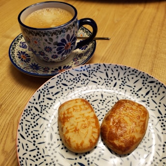 A decorated ceramic cup filled with frothy coffee is placed on a matching saucer, both featuring intricate floral patterns. In front of the cup, a plate with a blue and white design holds two golden-brown pastries. The setup rests on a wooden surface.