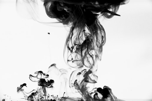 Black ink forms intricate, swirling patterns as it disperses in water against a stark white background. The image captures the fluid motion of the ink creating abstract shapes with varying opacity.