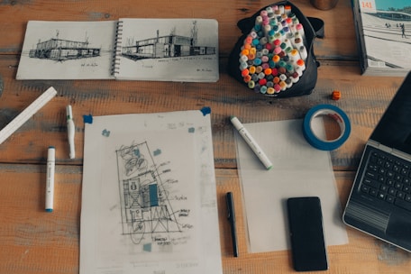 An assortment of architectural sketches and drafting tools arranged on a wooden table. A notebook with architectural designs, various markers, and measuring tools are visible. A black pouch filled with color markers, a roll of blue tape, and a small orange die are also present. A laptop and a smartphone are neatly placed, suggesting a working environment.