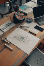 A wooden table scattered with various art supplies and electronics. There are colorful markers in a holder, a roll of blue tape, sketch papers with drawings and notes, and a laptop slightly open. The scene suggests a creative workspace with an active project in progress.