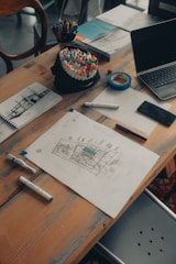 A wooden table scattered with various art supplies and electronics. There are colorful markers in a holder, a roll of blue tape, sketch papers with drawings and notes, and a laptop slightly open. The scene suggests a creative workspace with an active project in progress.