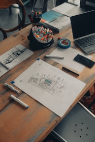 A wooden table scattered with various art supplies and electronics. There are colorful markers in a holder, a roll of blue tape, sketch papers with drawings and notes, and a laptop slightly open. The scene suggests a creative workspace with an active project in progress.