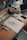 A wooden table scattered with various art supplies and electronics. There are colorful markers in a holder, a roll of blue tape, sketch papers with drawings and notes, and a laptop slightly open. The scene suggests a creative workspace with an active project in progress.