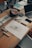 A wooden table scattered with various art supplies and electronics. There are colorful markers in a holder, a roll of blue tape, sketch papers with drawings and notes, and a laptop slightly open. The scene suggests a creative workspace with an active project in progress.