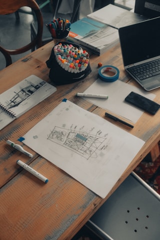 A wooden table scattered with various art supplies and electronics. There are colorful markers in a holder, a roll of blue tape, sketch papers with drawings and notes, and a laptop slightly open. The scene suggests a creative workspace with an active project in progress.
