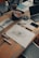 A wooden table scattered with various art supplies and electronics. There are colorful markers in a holder, a roll of blue tape, sketch papers with drawings and notes, and a laptop slightly open. The scene suggests a creative workspace with an active project in progress.