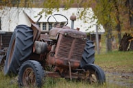 A rusty, vintage tractor is parked on grass, with large worn tires and a weathered body. In the background, there is a farmhouse with a white roof partially visible through sparse trees with autumn leaves.
