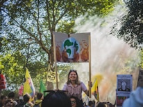 A crowd gathers for a protest in a park-like setting, with trees providing a natural backdrop. Many people hold signs, including one prominently displaying an illustration of Earth on fire, signaling climate change concerns. Sunlight filters through the trees, creating a serene yet determined atmosphere.