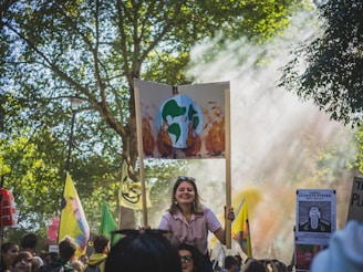 A crowd gathers for a protest in a park-like setting, with trees providing a natural backdrop. Many people hold signs, including one prominently displaying an illustration of Earth on fire, signaling climate change concerns. Sunlight filters through the trees, creating a serene yet determined atmosphere.