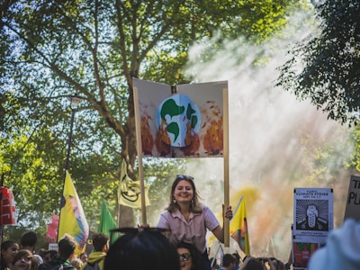 A crowd gathers for a protest in a park-like setting, with trees providing a natural backdrop. Many people hold signs, including one prominently displaying an illustration of Earth on fire, signaling climate change concerns. Sunlight filters through the trees, creating a serene yet determined atmosphere.
