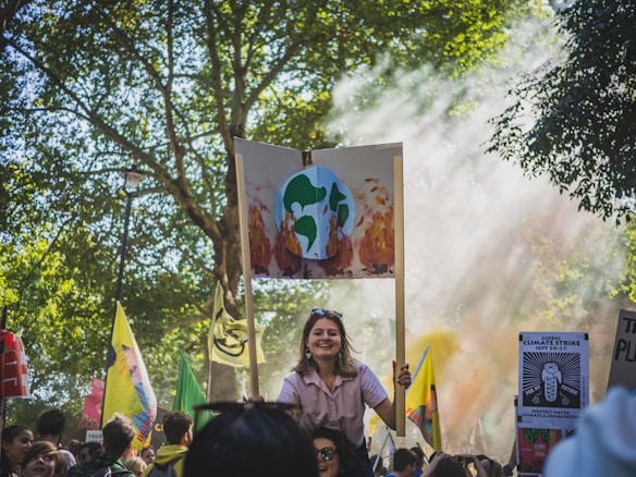 A crowd gathers for a protest in a park-like setting, with trees providing a natural backdrop. Many people hold signs, including one prominently displaying an illustration of Earth on fire, signaling climate change concerns. Sunlight filters through the trees, creating a serene yet determined atmosphere.