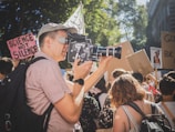 A person is filming a crowd of protesters using an old-fashioned film camera. Many people hold signs and banners, some of which have slogans like 'Science Not Silence'. The scene is outdoors, with sunlight filtering through trees, creating patches of light and shadow.