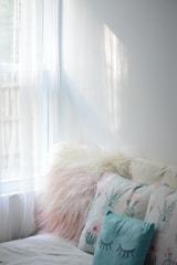 Sunlight filters through sheer white curtains, casting soft shadows in a cozy corner. Plush pillows with various textures and patterns, including a fluffy faux fur and decorative cactus designs, rest on a light-colored sofa.