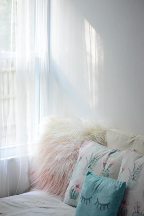 Warm sunlight filtering through sheer curtains onto a cozy reading nook with soft nude and white cushions.