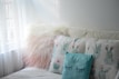 A cozy seating area features a fluffy, pinkish-white pillow beside two pillows with pastel cactus designs. The light blue pillow in front has closed eyes drawn on it, contributing to the calming atmosphere created by soft natural light coming through sheer curtains.