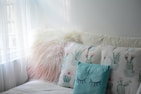 A cozy seating area features a fluffy, pinkish-white pillow beside two pillows with pastel cactus designs. The light blue pillow in front has closed eyes drawn on it, contributing to the calming atmosphere created by soft natural light coming through sheer curtains.