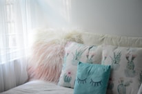 A cozy seating area features a fluffy, pinkish-white pillow beside two pillows with pastel cactus designs. The light blue pillow in front has closed eyes drawn on it, contributing to the calming atmosphere created by soft natural light coming through sheer curtains.