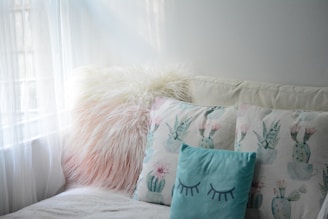 A cozy seating area features a fluffy, pinkish-white pillow beside two pillows with pastel cactus designs. The light blue pillow in front has closed eyes drawn on it, contributing to the calming atmosphere created by soft natural light coming through sheer curtains.