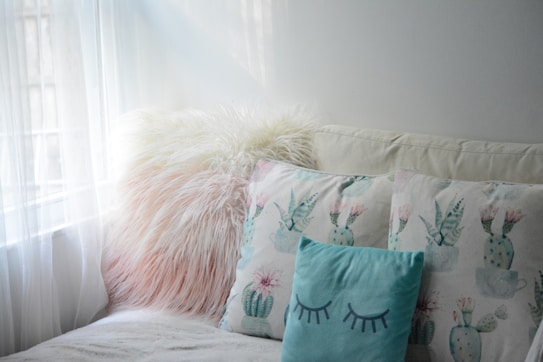 A cozy seating area features a fluffy, pinkish-white pillow beside two pillows with pastel cactus designs. The light blue pillow in front has closed eyes drawn on it, contributing to the calming atmosphere created by soft natural light coming through sheer curtains.