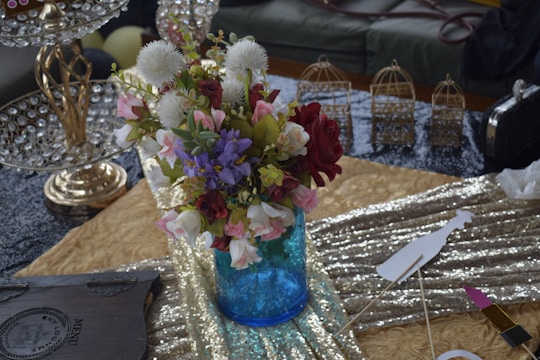 A vibrant bouquet of flowers arranged in a bright blue vase with a mix of red, pink, white, and purple blossoms, along with green foliage. The background includes a textured golden tablecloth, a metallic ornament with crystal detailing, and several small decorative birdcages. A shimmering sequin fabric adds to the luxurious setting.