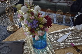 A vibrant bouquet of flowers arranged in a bright blue vase with a mix of red, pink, white, and purple blossoms, along with green foliage. The background includes a textured golden tablecloth, a metallic ornament with crystal detailing, and several small decorative birdcages. A shimmering sequin fabric adds to the luxurious setting.