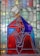 A stained glass artwork featuring a figure adorned in blue and red robes, with detailed geometric patterns. The figure has a serene expression, positioned in a praying gesture, and is crowned. Various hues of green, yellow, and blue form the background, enhancing the composition with a celestial halo effect.