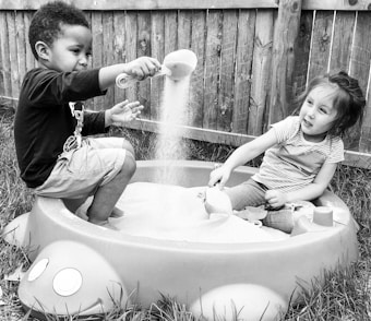 Two young children are playing happily in a sandbox that is designed to resemble a ladybug. One child is pouring sand from a scoop, and the other is seated, engaging in the activity. A wooden fence forms the background, and grass surrounds the sandbox.